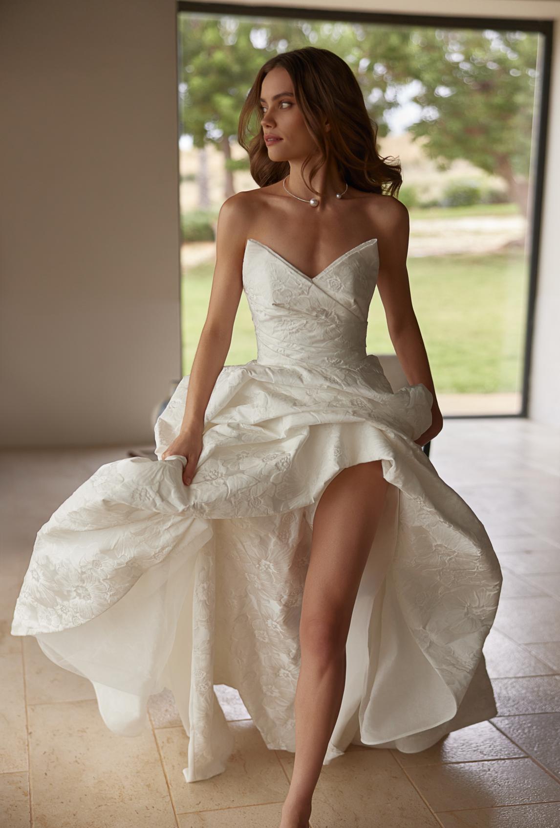 Bride in a lush lace wedding dress with an open bodice and a long train outdoors among greenery.